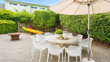 A round outdoor dining table with white chairs, a yellow kayak, and a large umbrella in a garden area.