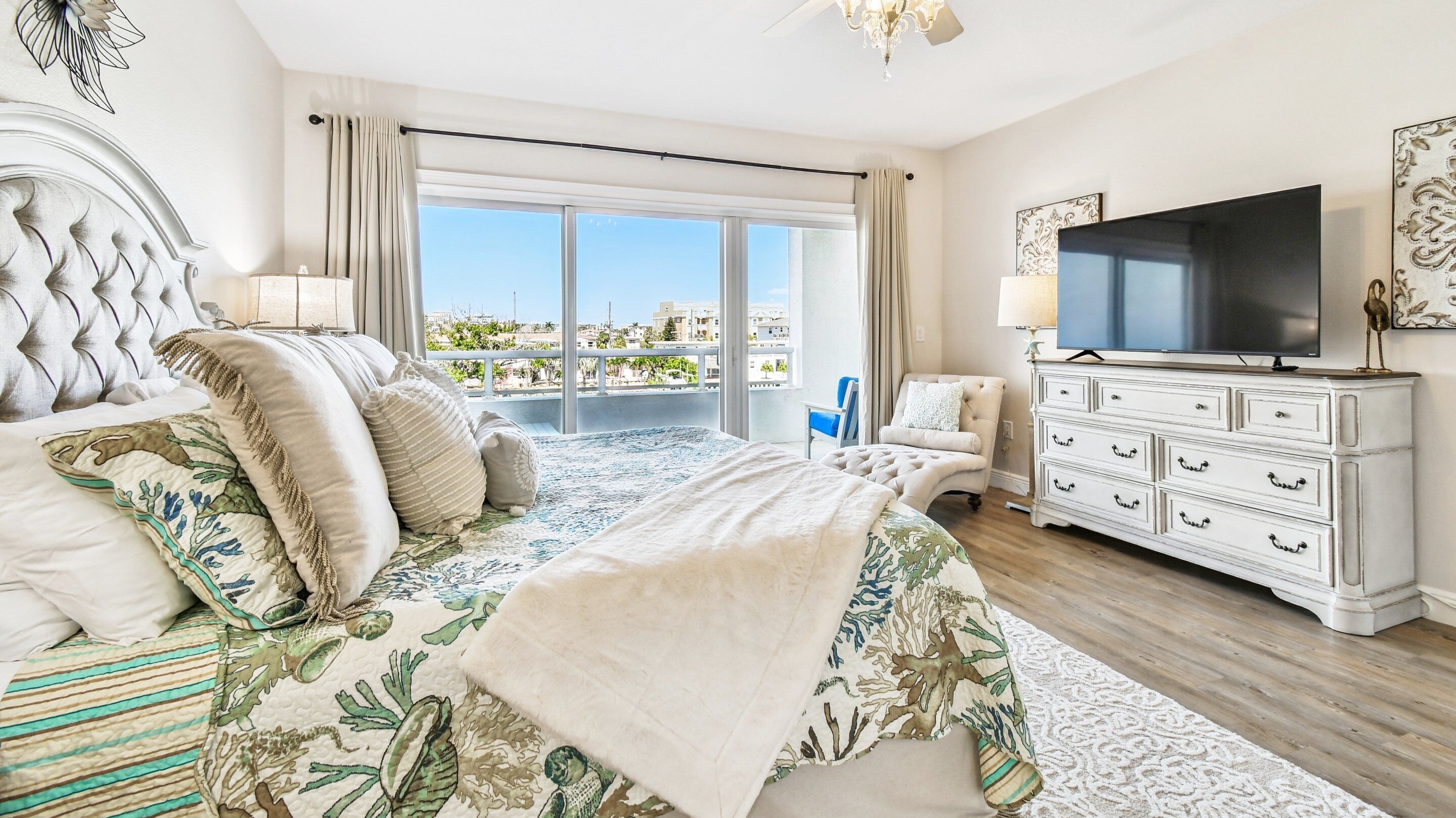A bedroom with a large bed, a TV, a dresser, and a balcony with a city view.