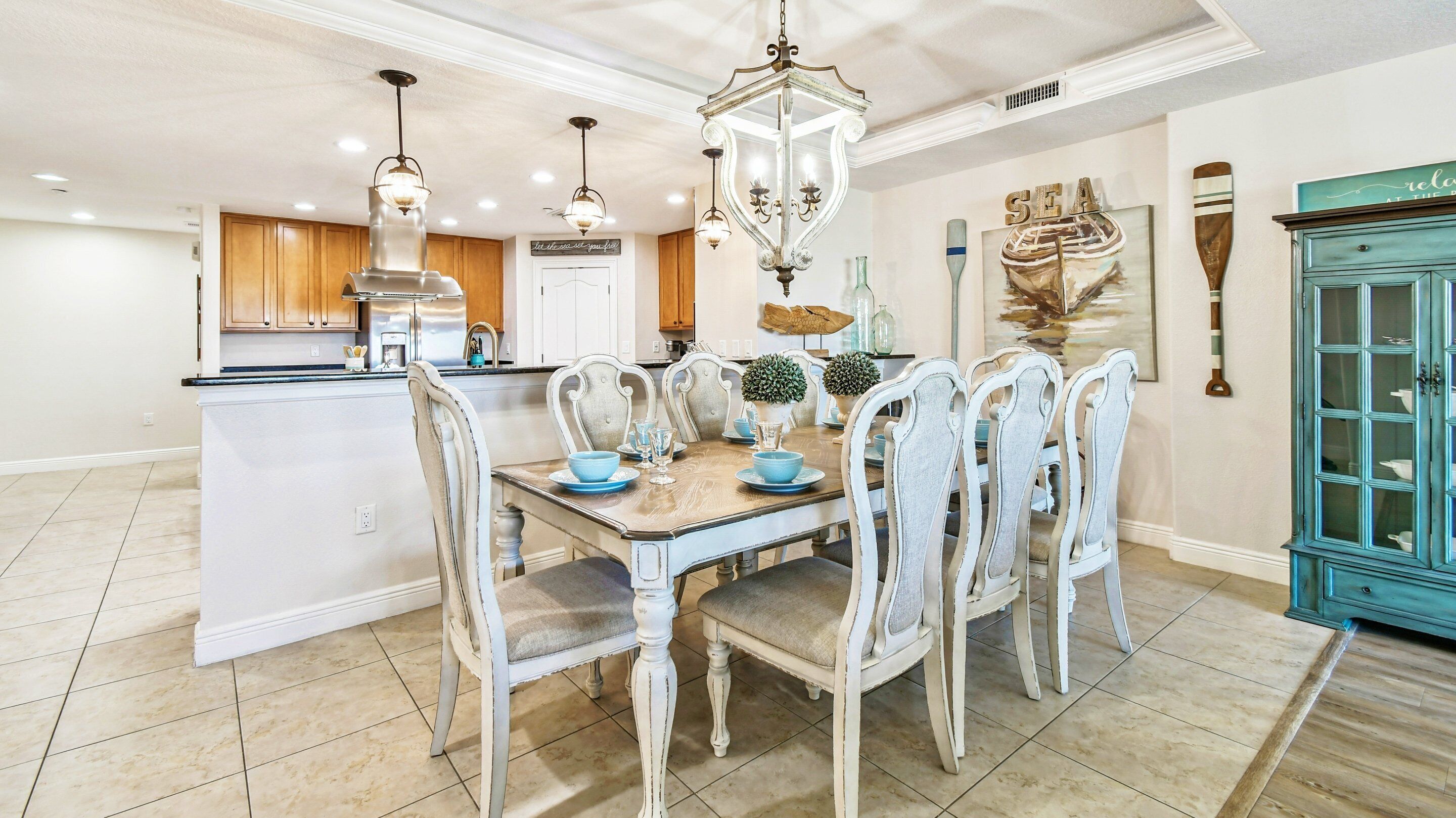 A dining area with a wooden table and chairs, a chandelier, and a kitchen in the background.