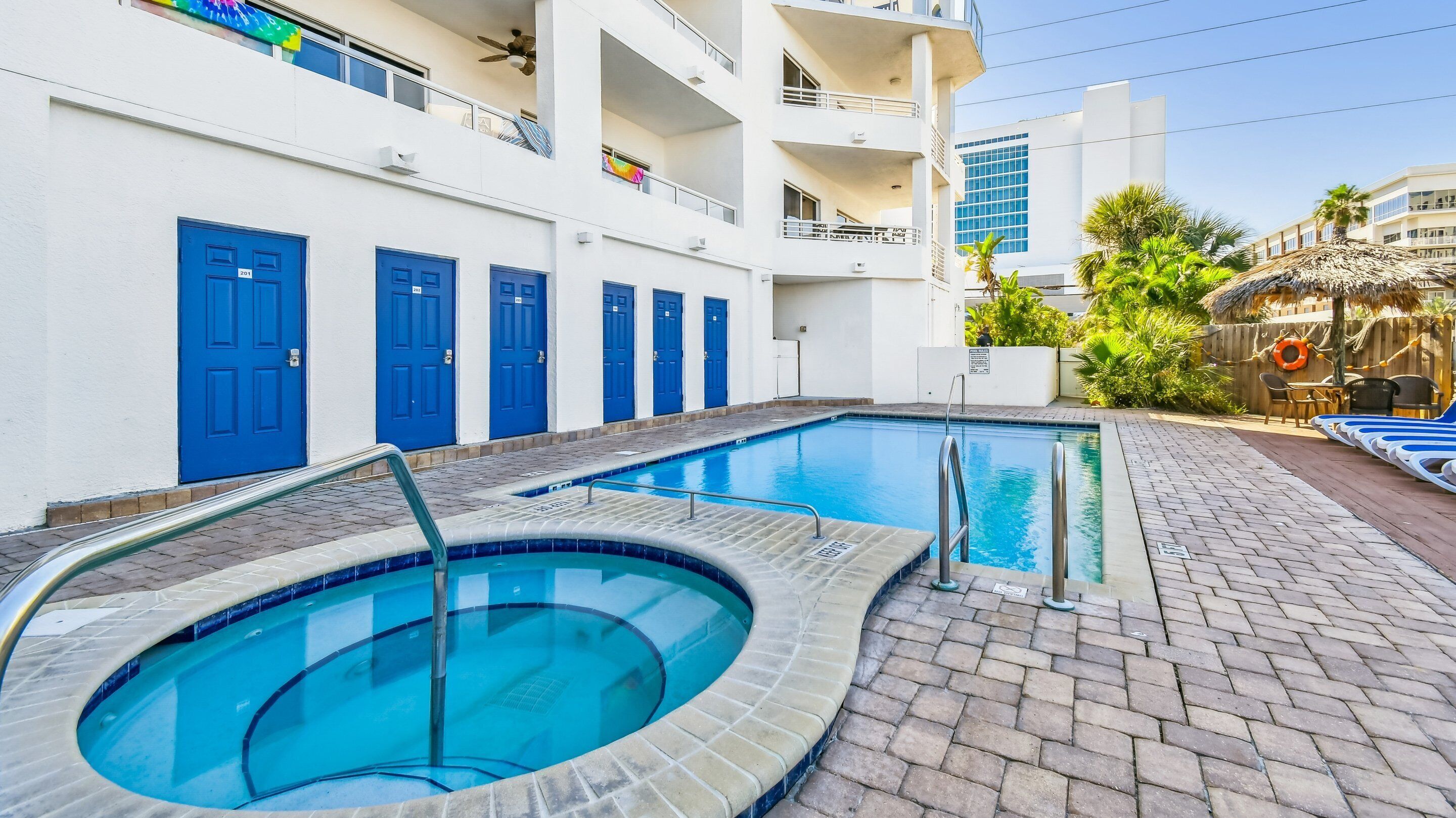 A swimming pool with a hot tub, surrounded by a brick patio and a white building with blue doors.