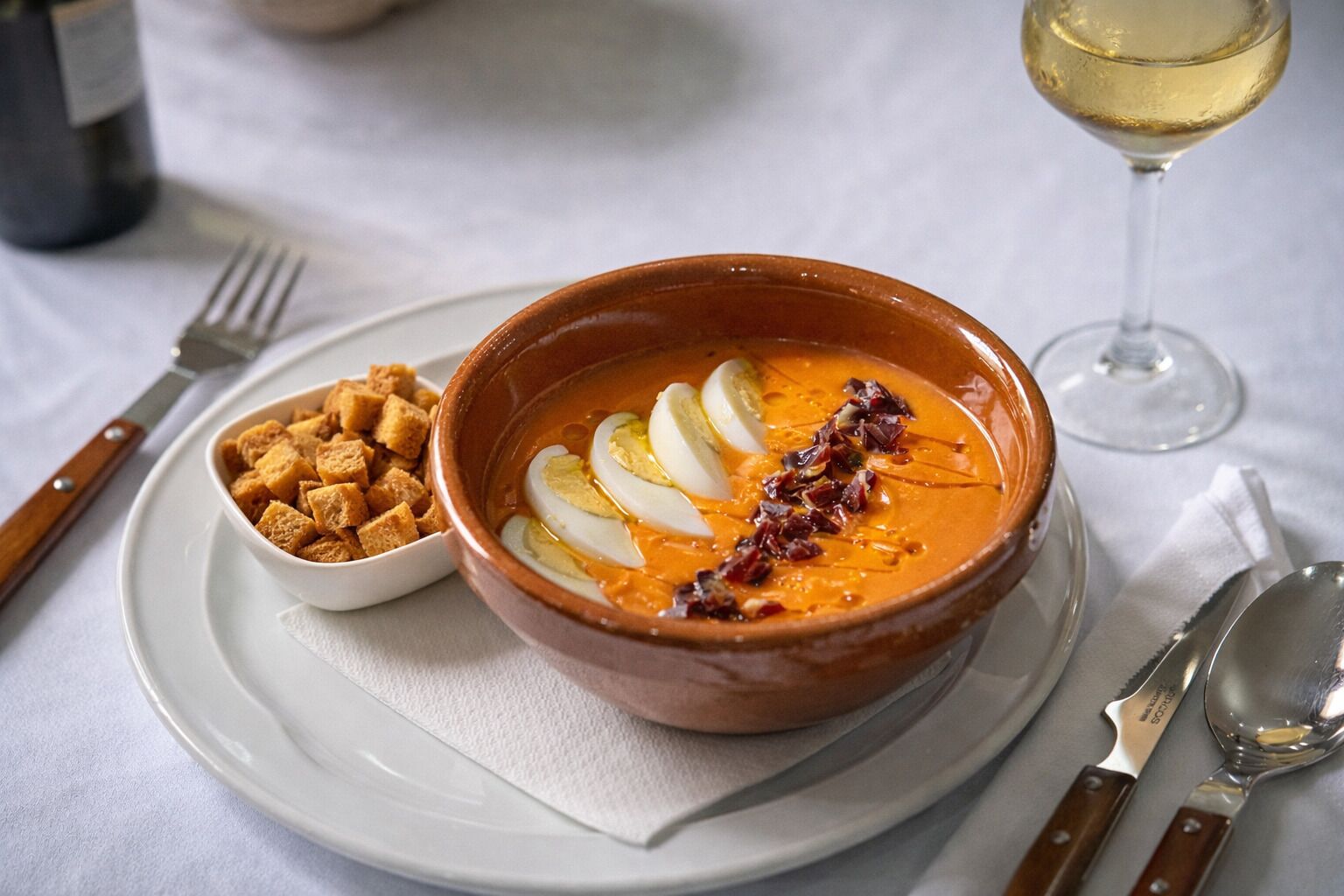 Un bol de soupe garni de tranches d’œuf et de croûtons, servi sur une assiette blanche, accompagné d’un verre de vin blanc et de couverts.