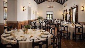A spacious dining hall with round tables set for multiple guests, featuring chandeliers and wall-mounted lighting.