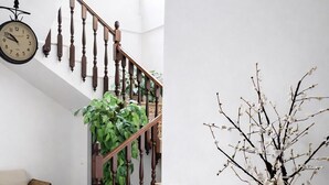 A staircase with wooden balusters, a clock, a wicker sofa, a potted plant, and a tall vase with branches.