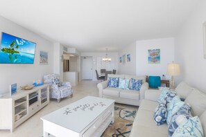 Un salon moderne avec un canapé blanc, un fauteuil à motifs et une table basse. Un téléviseur à écran plat est fixé au mur, un lustre est suspendu au plafond et, au fond, on aperçoit un coin repas.