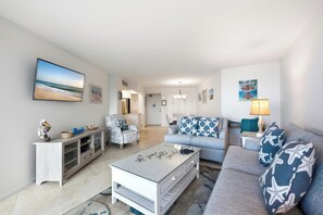 A spacious living room with a sofa, armchair, and coffee table. A television is mounted on the wall, and there is a door leading to another room.