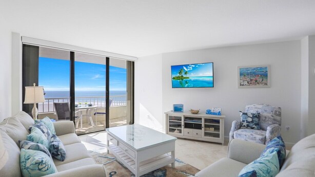 A modern living room with a view of the ocean, a white sofa, a coffee table, and a TV on the wall.