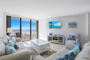 Un salón moderno con vistas al océano, un sofá blanco, una mesa de centro y un televisor en la pared.