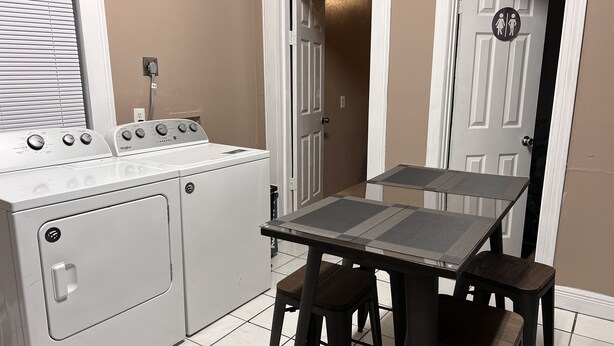 Une cuisine compacte équipée d’un lave-linge et d’un sèche-linge, d’une petite table avec des tabourets et d’une porte ornée d’un symbole indiquant le genre.