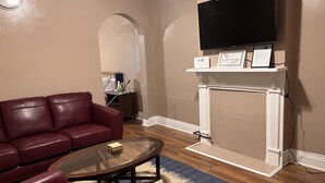Un salon avec un canapé en cuir rouge, une table basse en verre, une cheminée sur laquelle est fixée une télévision, et une petite étagère avec des panneaux décoratifs.