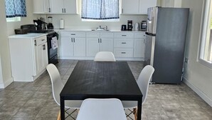 Een kleine keuken met witte kastjes, een zwart aanrecht en een grijze koelkast. In het midden staat een eettafel met vier witte stoelen.