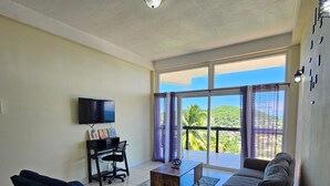 Een woonkamer met een bank, een salontafel, een televisie en een balkon met uitzicht op groen.
