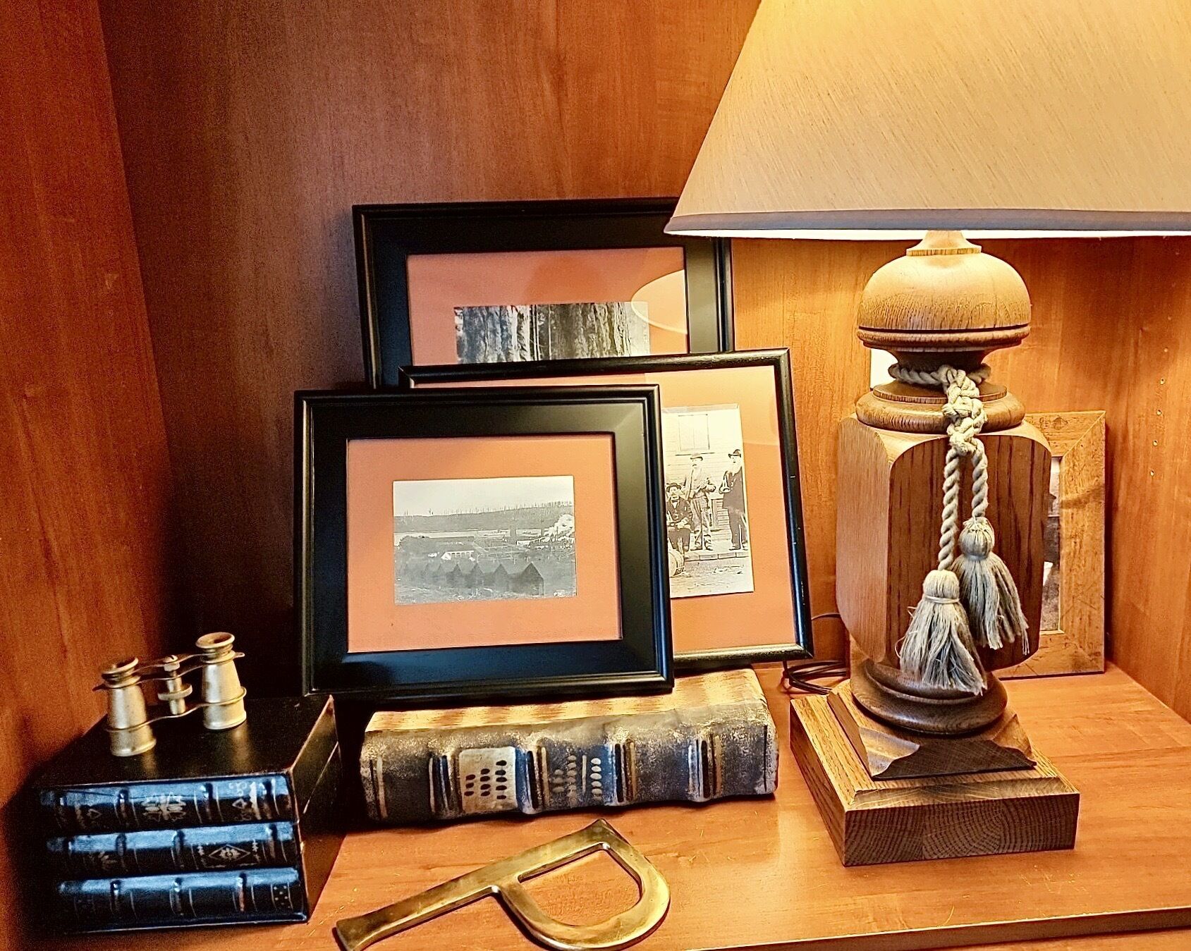 A wooden desk with framed photographs, a vintage lamp, a book, and a pair of glasses.