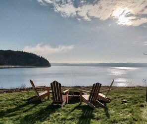 Deux fauteuils Adirondack sont placés face à un lac paisible, dont les rives sont bordées d’une forêt, sous un ciel nuageux.