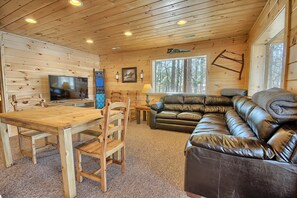 A cabin interior with a wooden dining table, chairs, a leather sofa, a television, and a large window with a forest view. - Lone Star Lodge - Gorgeous Lodge in Hocking Hills (Logan)