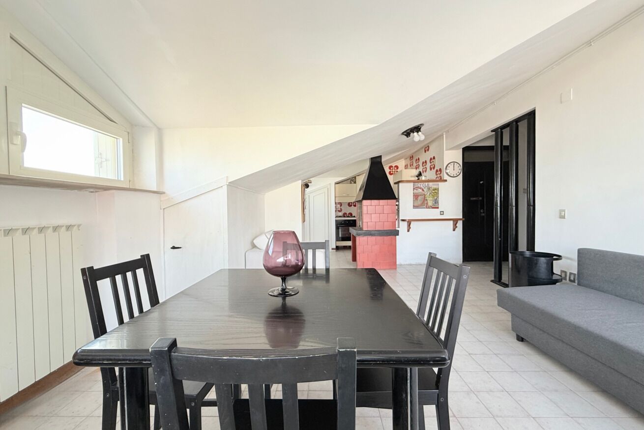 A dining area with a table, chairs, and a wine glass. There is a grey sofa and a kitchen area in the background.