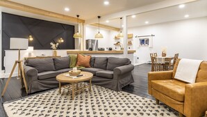 Un salon moderne avec un canapé gris, une table basse ronde et un fauteuil en cuir marron. On y trouve également un coin repas avec une table et des chaises, et une cuisine à l’arrière-plan.