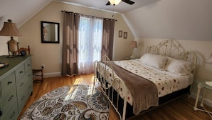 Une chambre à coucher comprenant un lit, un ventilateur de plafond, une table de chevet, une lampe, une commode, un miroir et une fenêtre avec des rideaux.