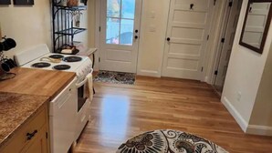 Une cuisine équipée d’une cuisinière, d’un four et d’un plan de travail. On y trouve une porte avec une fenêtre, un tapis et une étagère murale sur laquelle sont posés divers objets.