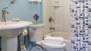 A bathroom with a sink, toilet, and bathtub. There is a towel rack with towels, a glass shelf with toiletries, and a shower curtain.