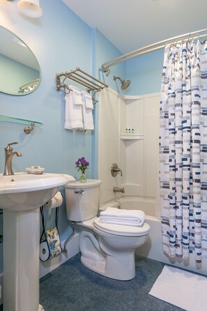 A bathroom with a sink, toilet, and bathtub. There is a towel rack with towels, a glass shelf with toiletries, and a shower curtain.