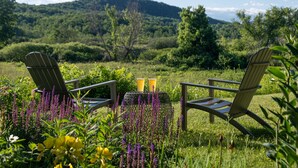 Dos sillas Adirondack mirando hacia una mesa con dos tazas, rodeadas de flores silvestres vibrantes y un paisaje verde exuberante con colinas al fondo.