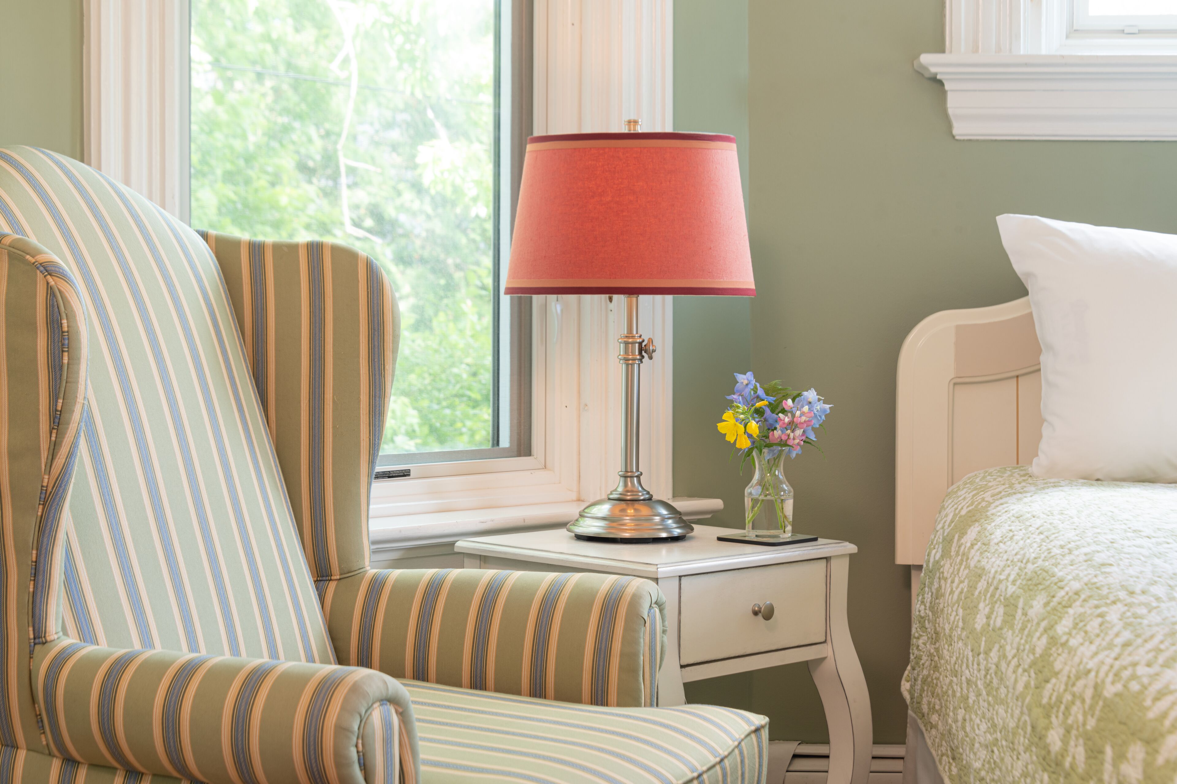 A room with a striped armchair, a bedside table with a lamp, a vase with flowers, and a bed with a green patterned bedspread.