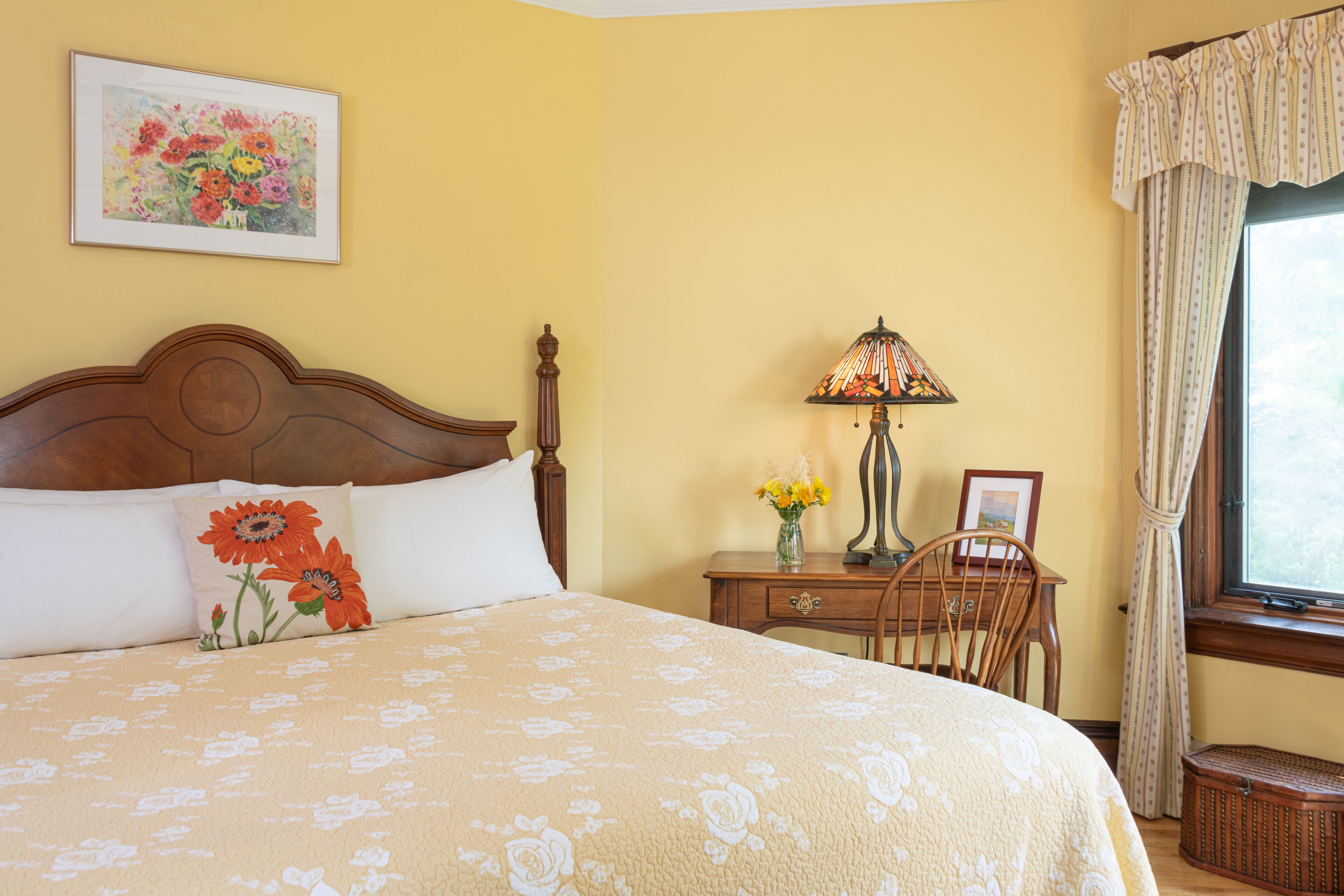 A bedroom with a large bed, a wooden nightstand, a lamp, a chair, and a window with curtains.