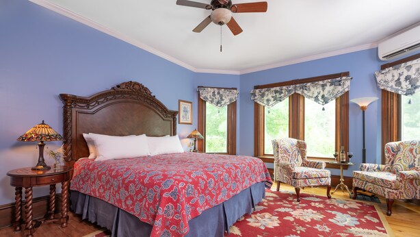 A bedroom with a large bed, two chairs, a ceiling fan, and a window with curtains.