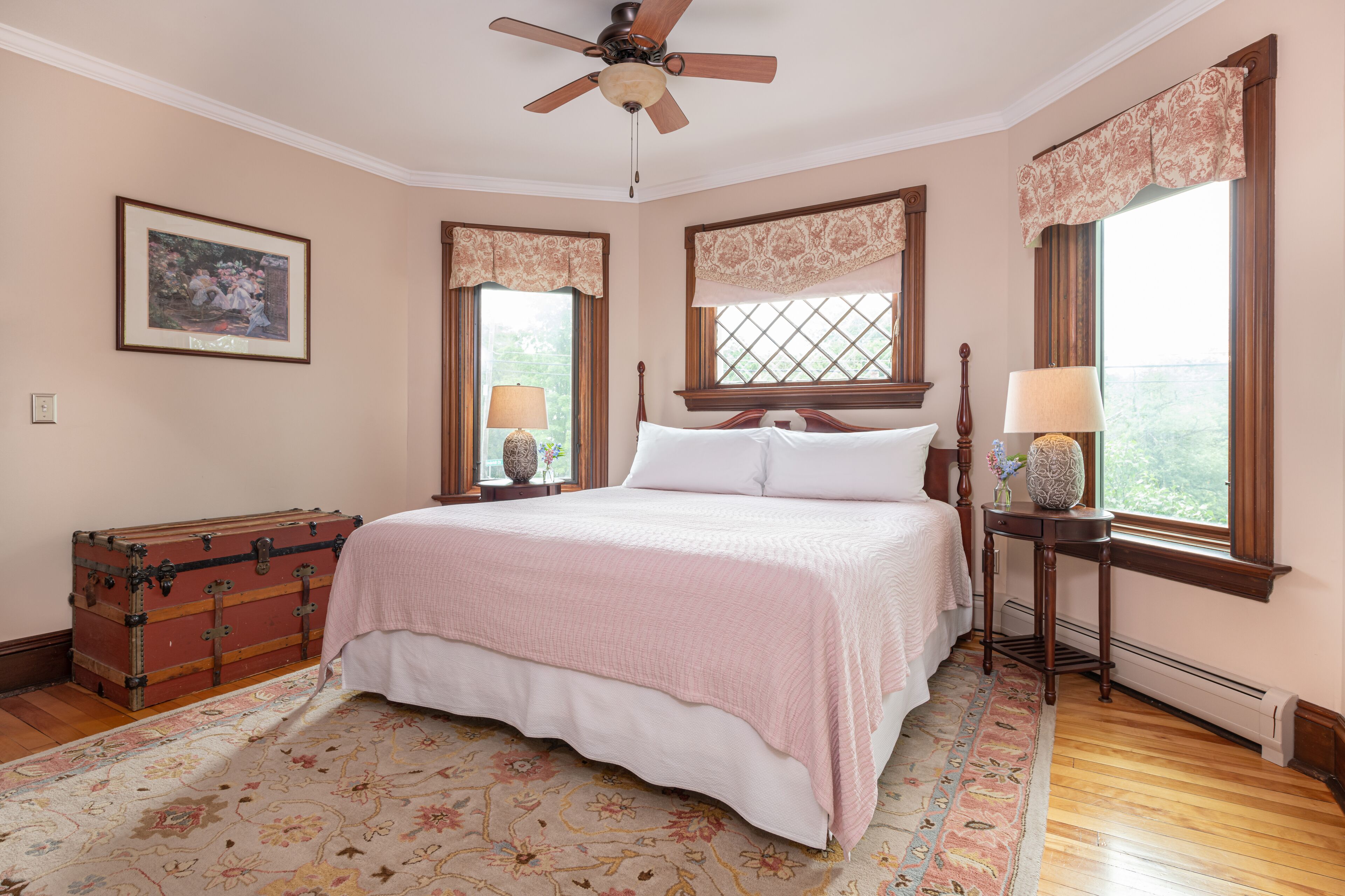 A bedroom with a large bed, a ceiling fan, a window with curtains, a bedside table with lamps, and a vintage trunk.
