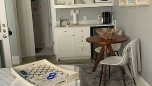 A compact room with a bed, a small table with a chair, a kitchen area with cabinets and a microwave, and a door leading to another room.