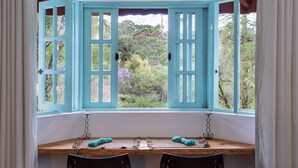 Une baie vitrée aux montants turquoise, une table en bois, deux chaises et une vue sur la verdure et les arbres.