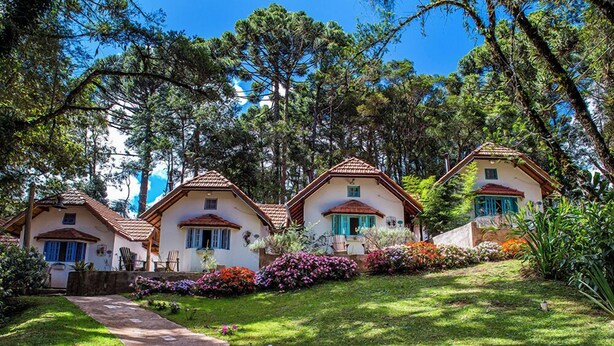 Une rangée de trois maisons blanches aux toits rouges et aux volets bleus, entourées d’une végétation luxuriante et sous un ciel bleu azur.