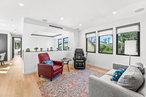 A modern living room with a red chair, a massage chair, and a grey sofa. There are large windows with views of greenery and a building. - Gorgeous home for Executive re-location, Digital Nomads, or Families (Huntington Beach)