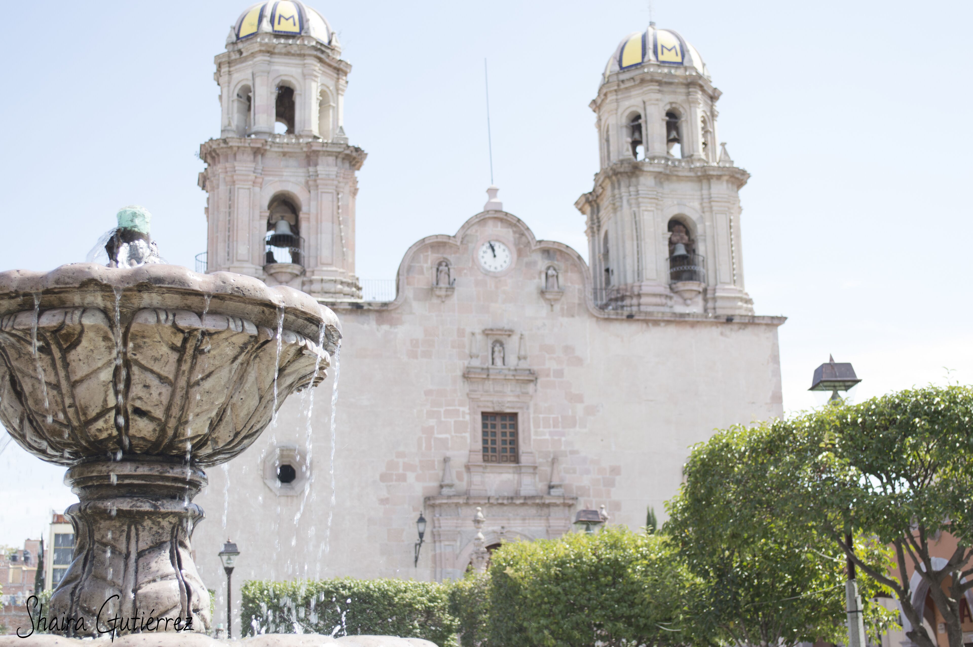 A historic building with two bell towers and a central dome, a fountain in the foreground, and a clear sky.