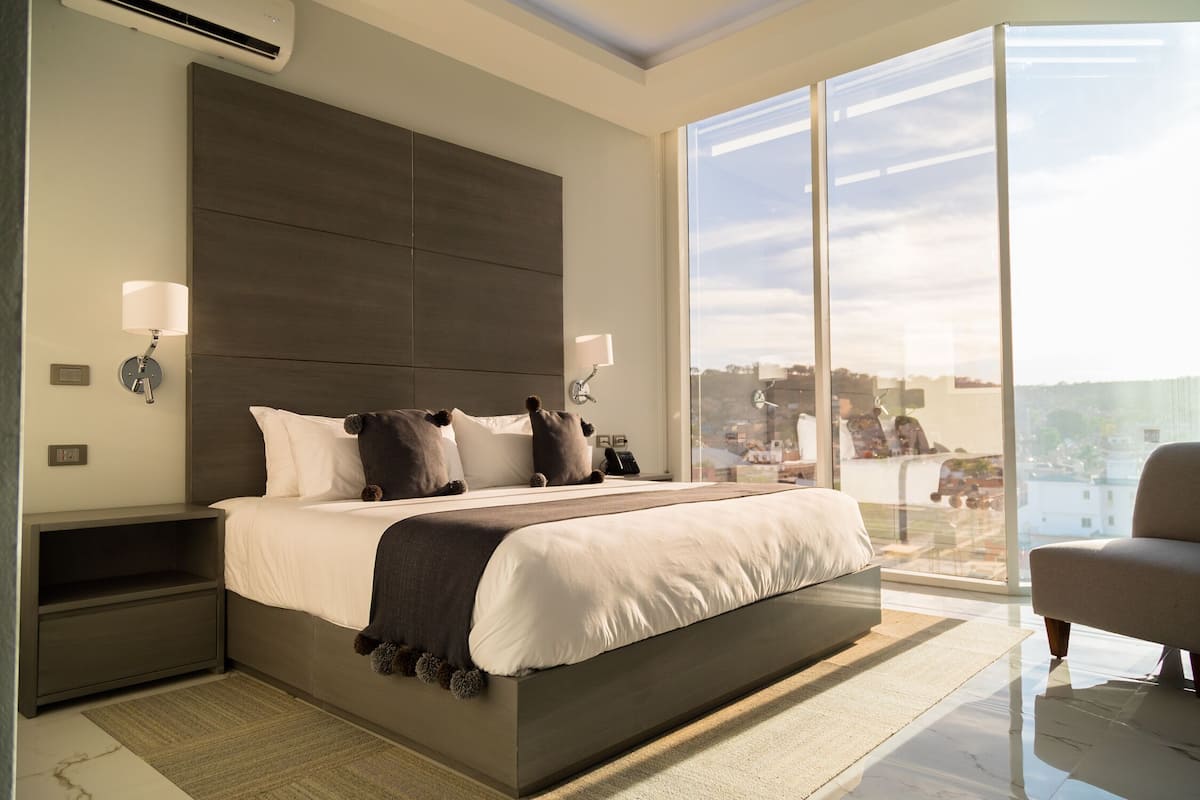 A modern bedroom with a large bed, bedside tables, a nightstand, a sofa, and a large window offering a city view.