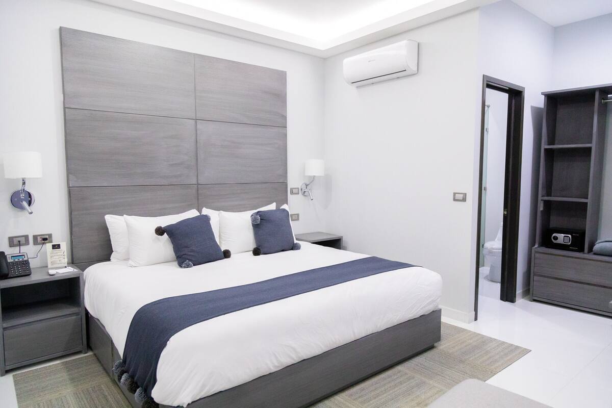 A modern hotel room with a large bed, bedside tables, a wardrobe, and an air conditioning unit.