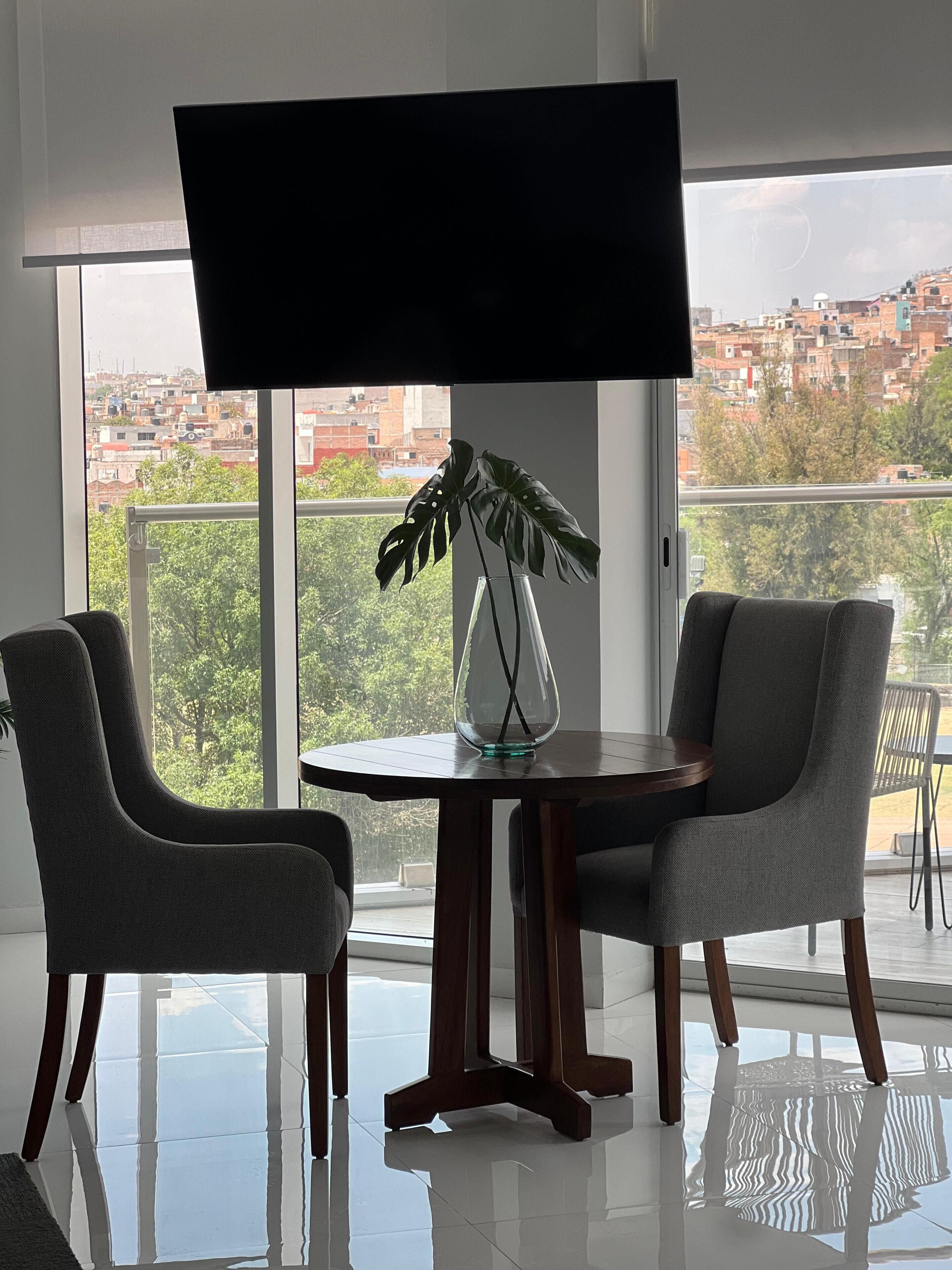 A modern living room with a large TV, two grey upholstered chairs, a glass vase with a plant, and a view of a cityscape through large windows.