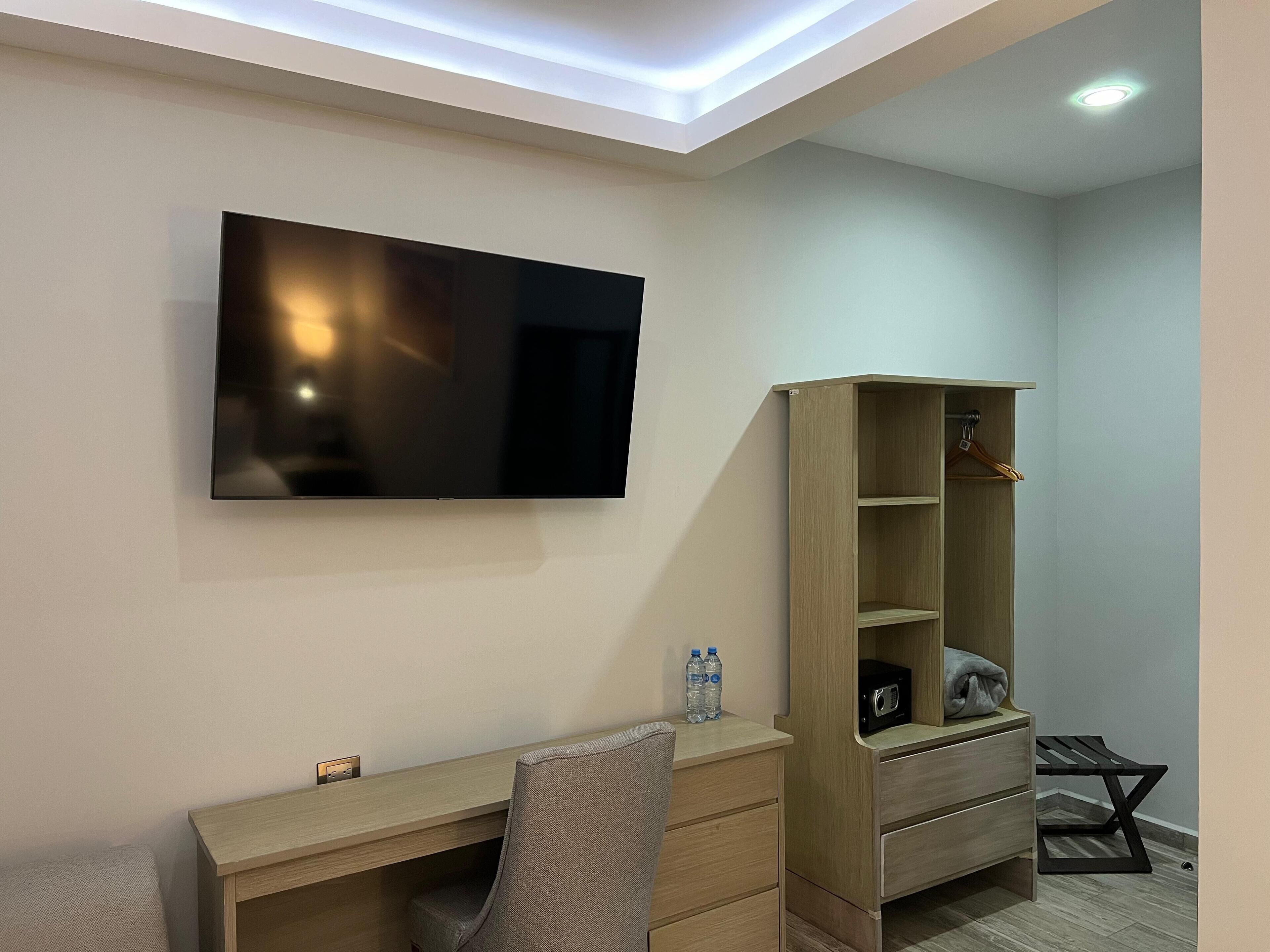 A modern room with a flat-screen TV mounted on the wall, a wooden desk with drawers, a chair, a wooden shelving unit, and a small table with a radio.