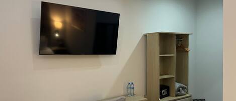 A modern room with a flat-screen TV mounted on the wall, a wooden desk with drawers, a chair, a wooden shelving unit, and a small table with a radio.