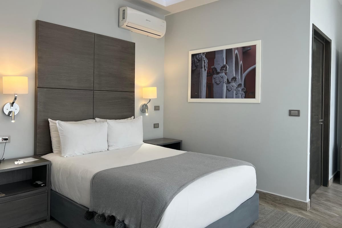 A modern hotel room with a large bed, two bedside tables, a wall-mounted air conditioner, and a framed artwork above the bed.
