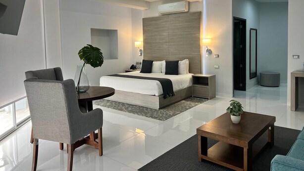 Une chambre d’hôtel moderne dotée d’un grand lit, de tables de chevet, d’un coin salon avec un fauteuil et une table, et d’une grande fenêtre.