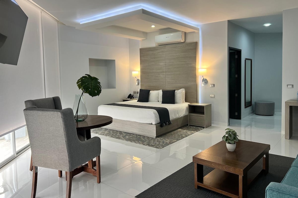 A modern hotel room with a large bed, bedside tables, a seating area with a chair and a table, and a large window.