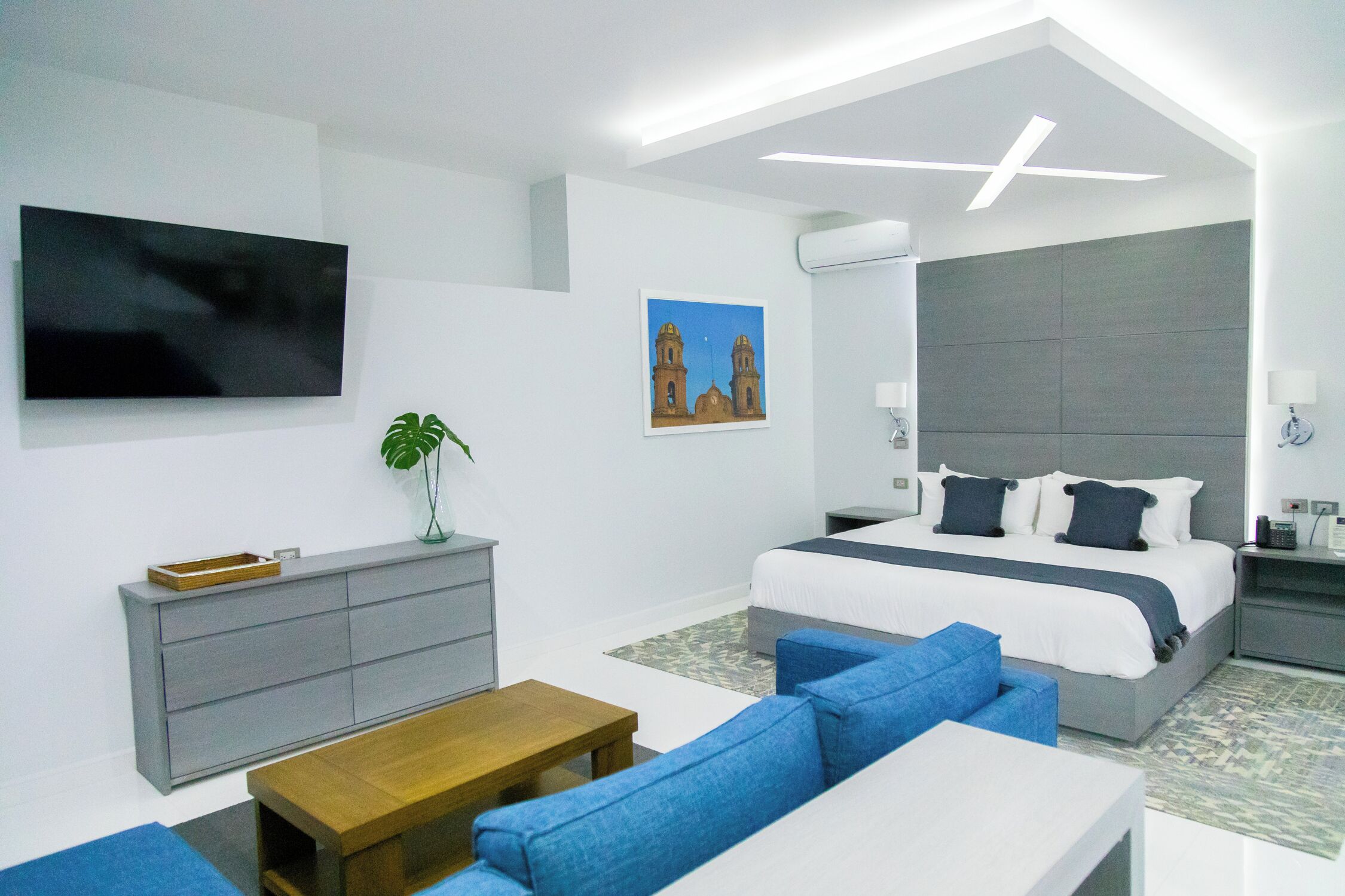 A modern hotel room with a large bed, a sofa, a TV, a desk, and a plant.
