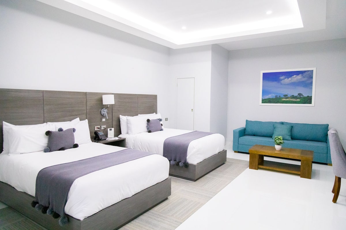 A modern hotel room with two beds, a sofa, a coffee table, and a painting on the wall.