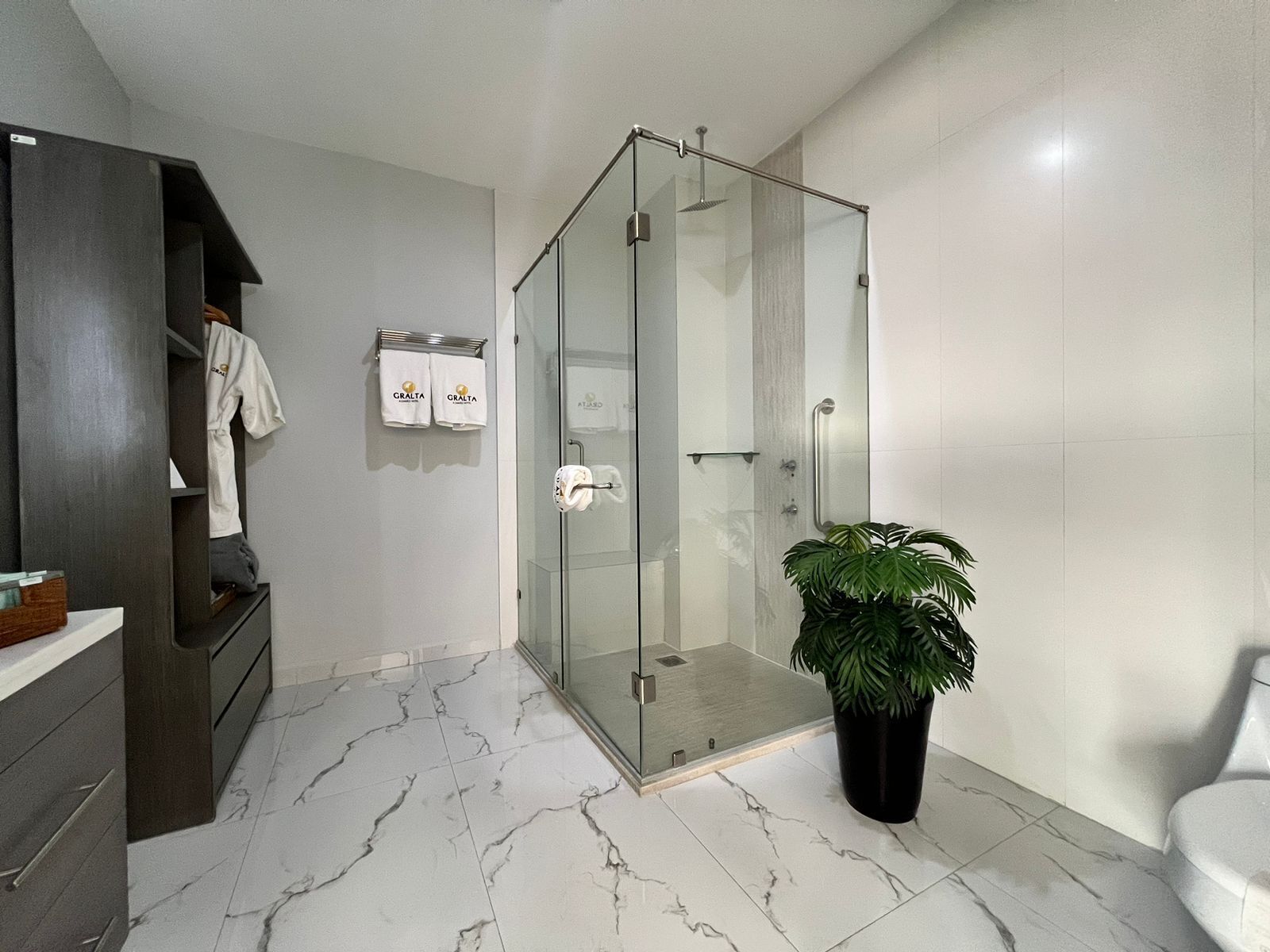 Un baño moderno con ducha de vidrio, piso de mármol, una planta en maceta y un perchero con toallas de la marca.