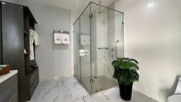 Un baño moderno con ducha de vidrio, piso de mármol, una planta en maceta y un perchero con toallas de la marca.
