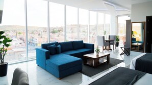 Un salon moderne avec un canapé d’angle bleu, une table basse en bois et une vue sur la ville grâce à de grandes fenêtres.