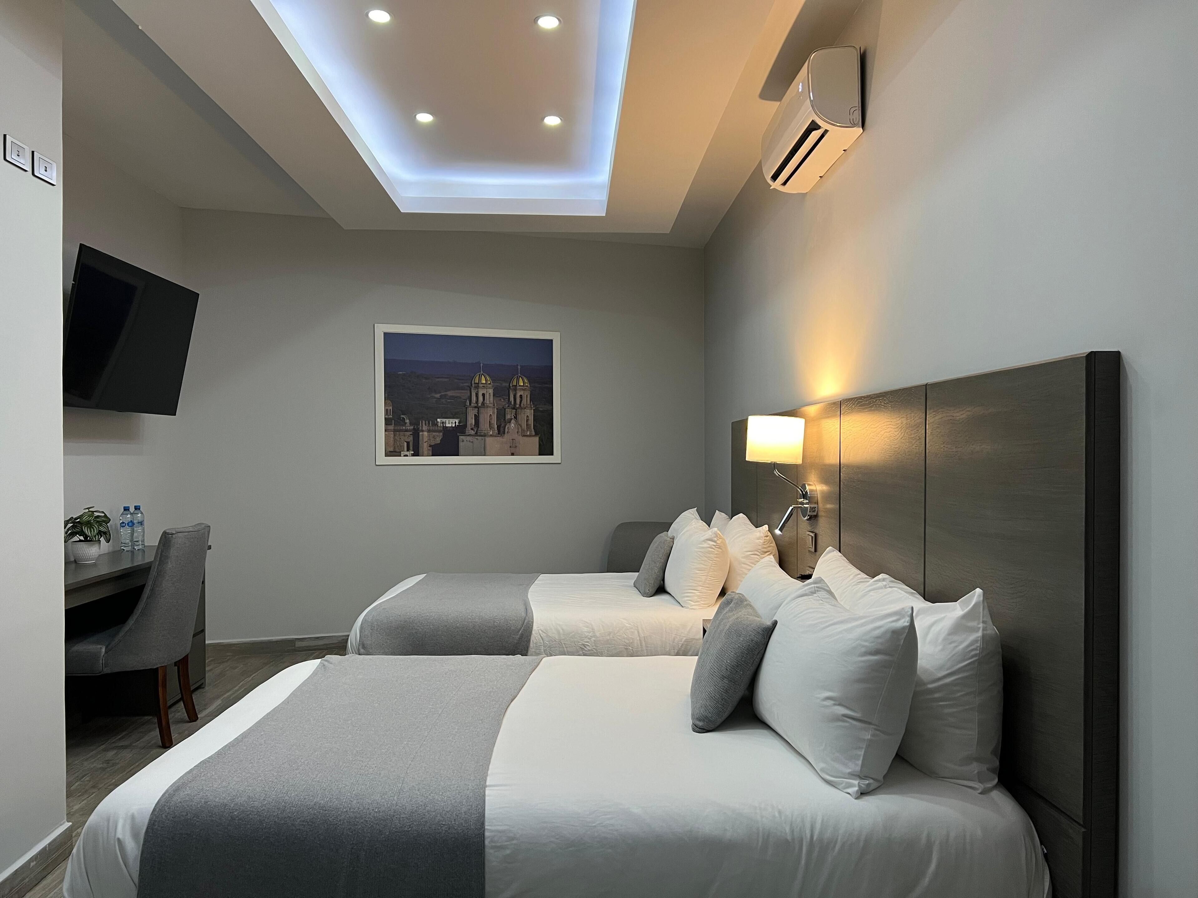 Une chambre d’hôtel avec deux lits, un bureau, une chaise, un téléviseur à écran plat, un tableau représentant un paysage urbain encadré et un appareil de climatisation.