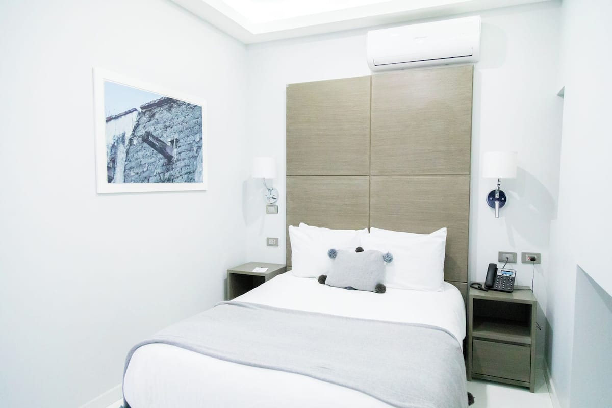 A hotel room with a bed, a nightstand with a phone, a wall-mounted air conditioner, and a framed picture on the wall.