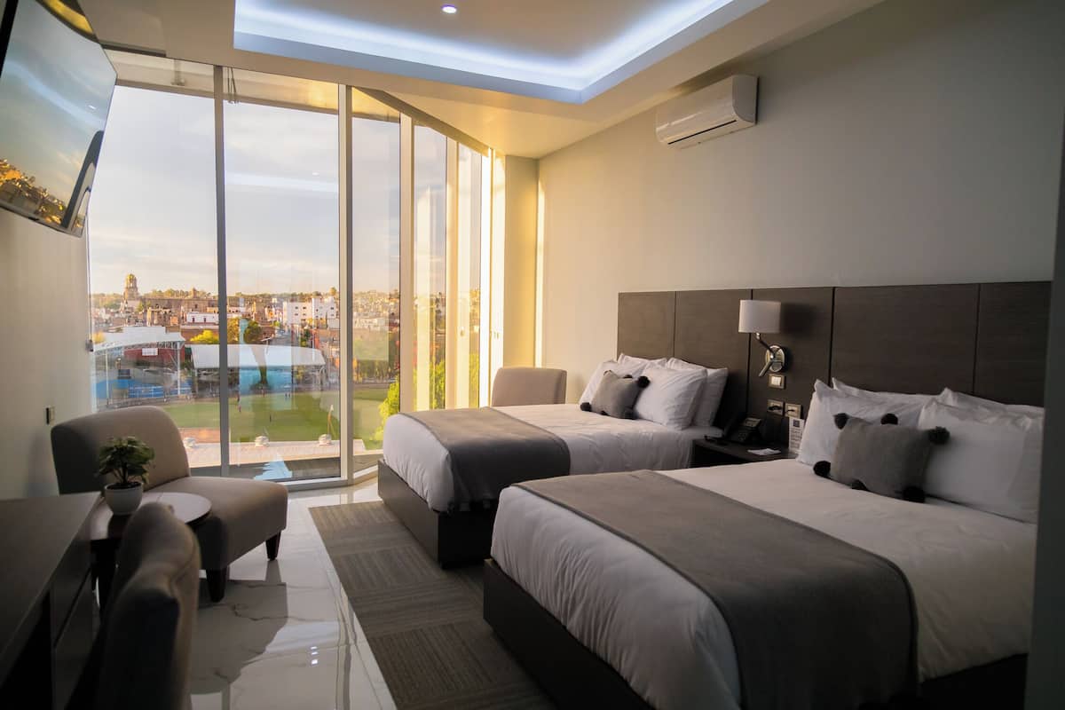 A modern hotel room with two beds, a sofa, a small table, and a large window offering a city view.